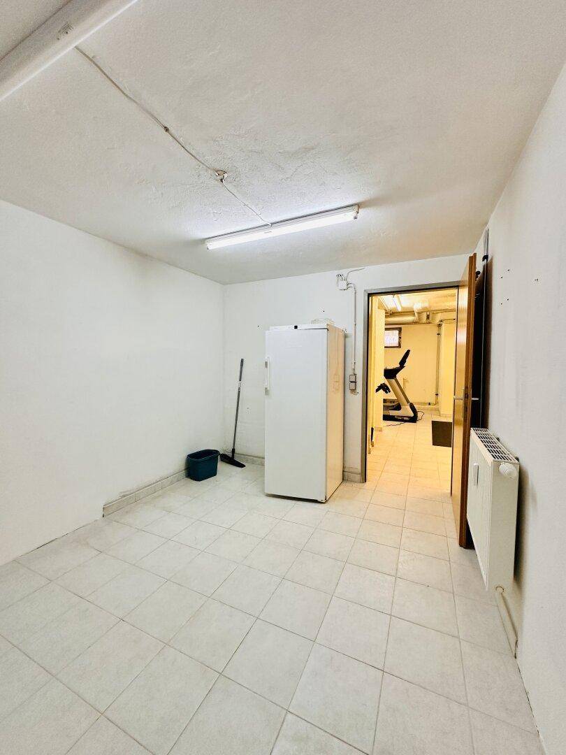 Heller Kellerraum mit Fliesenboden, Kühlschrank und Heizkörper, mit Blick auf einen Fitnessbereich.