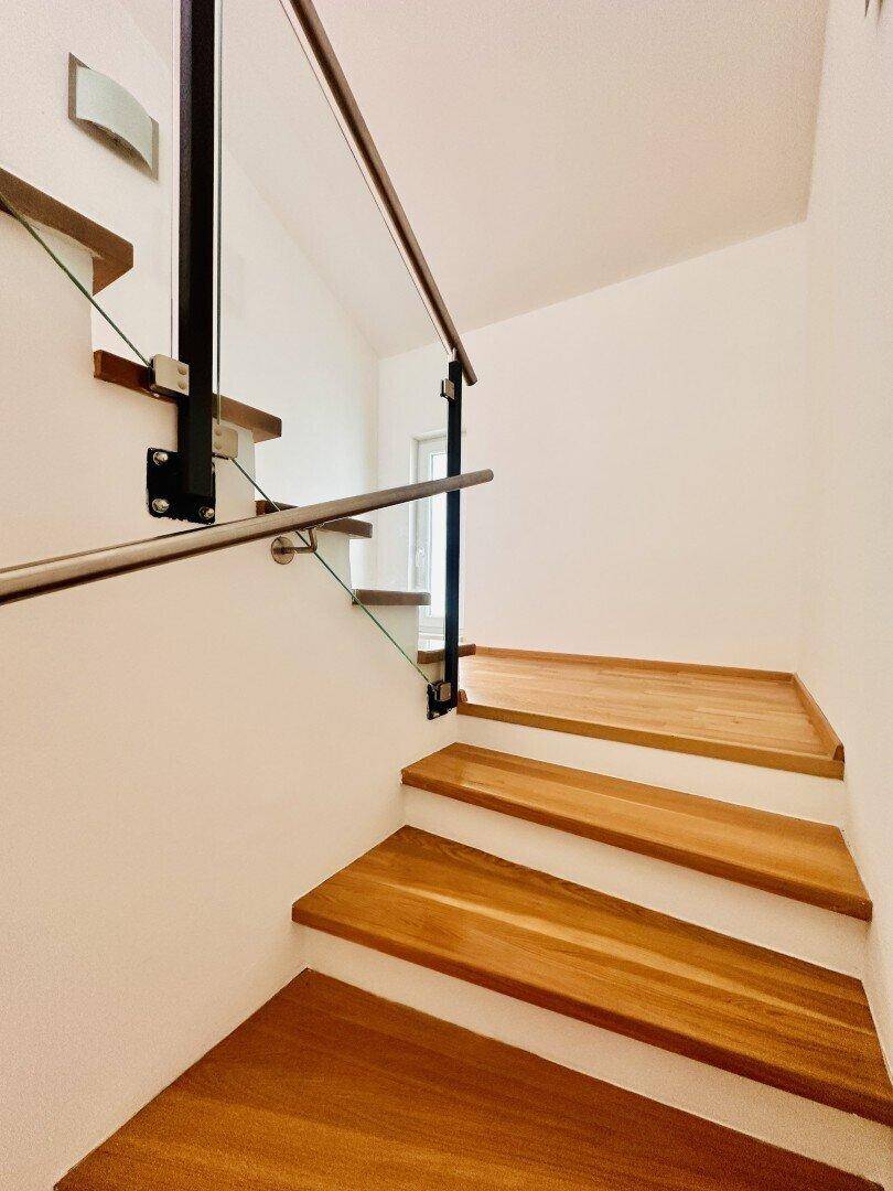 Moderne Holztreppe mit Glasgeländer und Edelstahlhandlauf in hellem Treppenhaus.