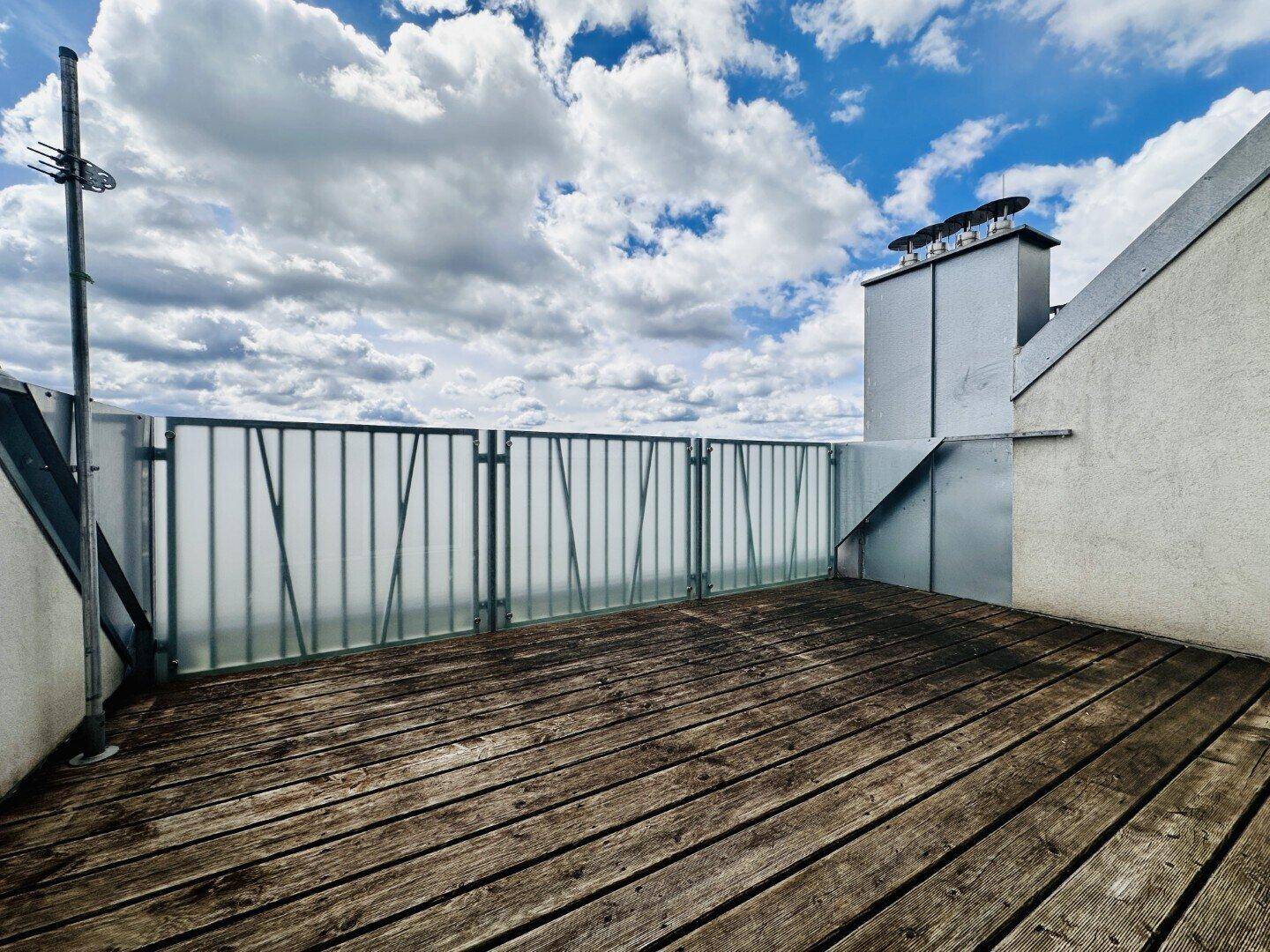 Geräumige Dachterrasse mit Holzboden und Metallgeländer unter blauem Himmel.