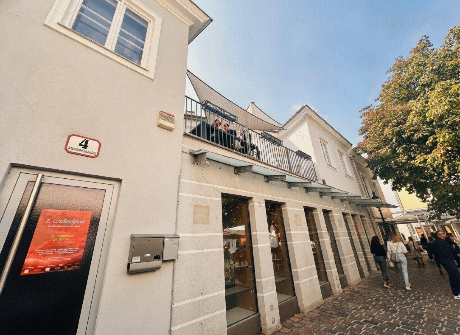 Ansicht der Gebäudefassade mit Geschäften im Erdgeschoss und einer zugänglichen Terrasse im Obergeschoss.