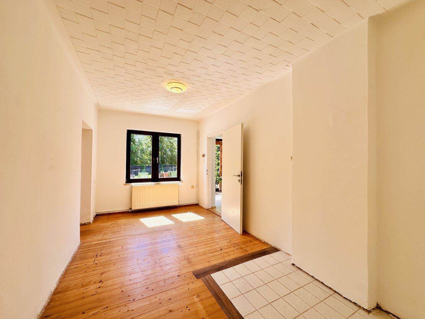 Helles Zimmer mit Holzplankenboden, Fenster und einem gefliesten Bereich am Eingang.