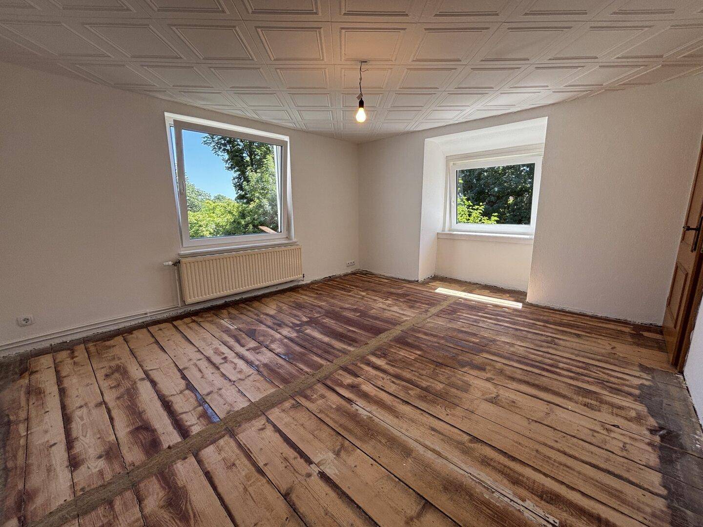 Helles Zimmer mit freigelegten Dielenböden, zwei Fenstern und einer strukturierten Decke.