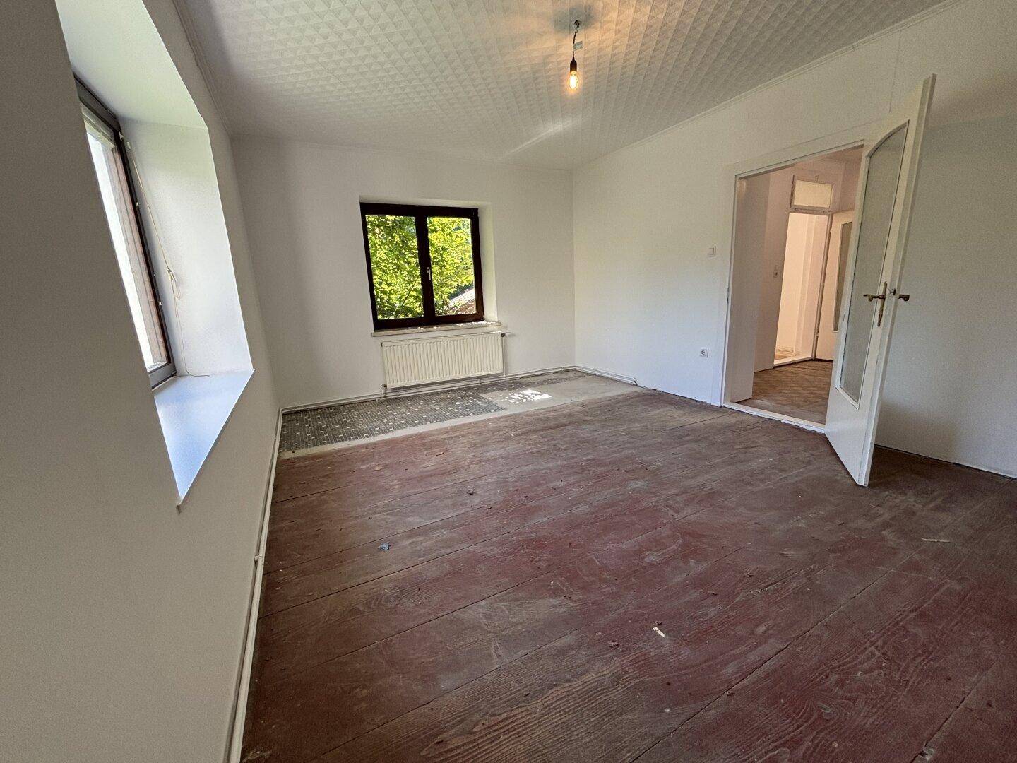 Helles Zimmer mit freigelegten Dielenböden, zwei Fenstern und Zugang zu einem weiteren Raum.