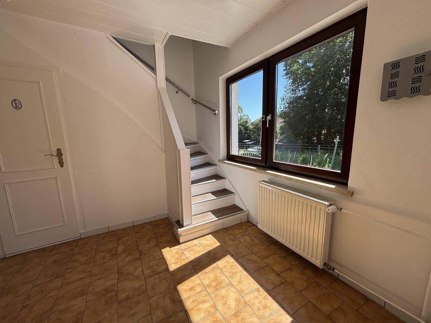 Heller Eingangsbereich mit gefliestem Boden, Treppe zum Obergeschoss und großem Fenster.