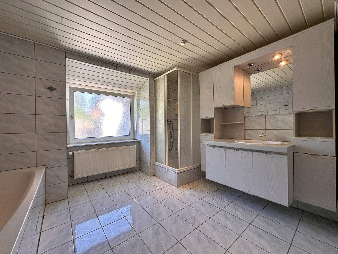 Helles Badezimmer mit Duschkabine, Waschtisch und Fenster, das Tageslicht hereinlässt.