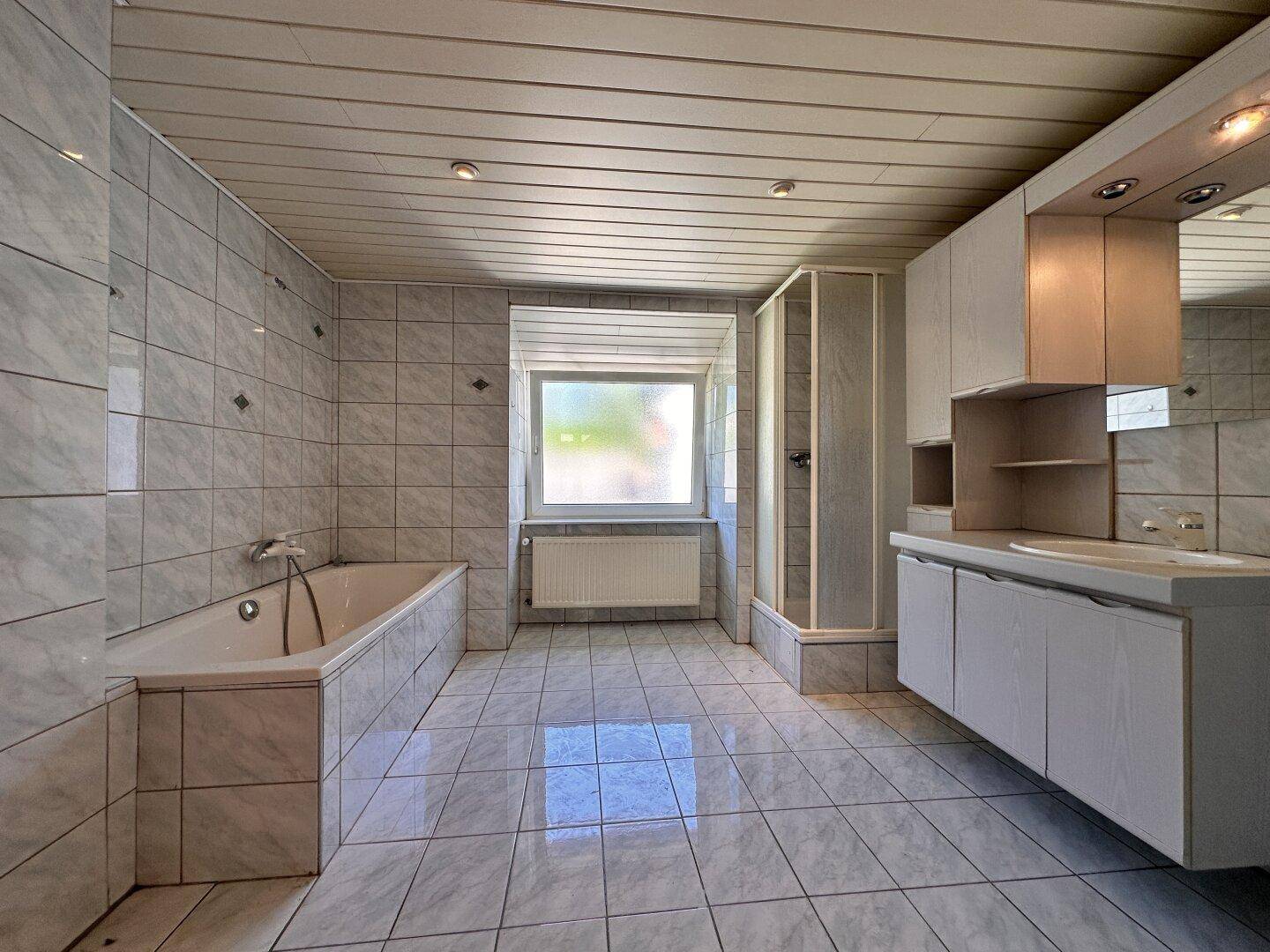 Geräumiges Badezimmer mit grauen Fliesen, Badewanne, Duschkabine und Waschtisch mit Spiegelschrank.