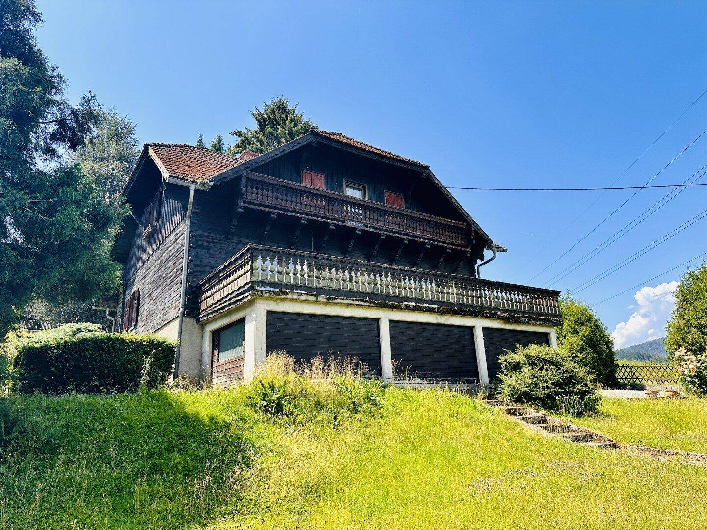 Traditionelles Holzhaus mit Balkon und Garagen, eingebettet in grüne Landschaft.