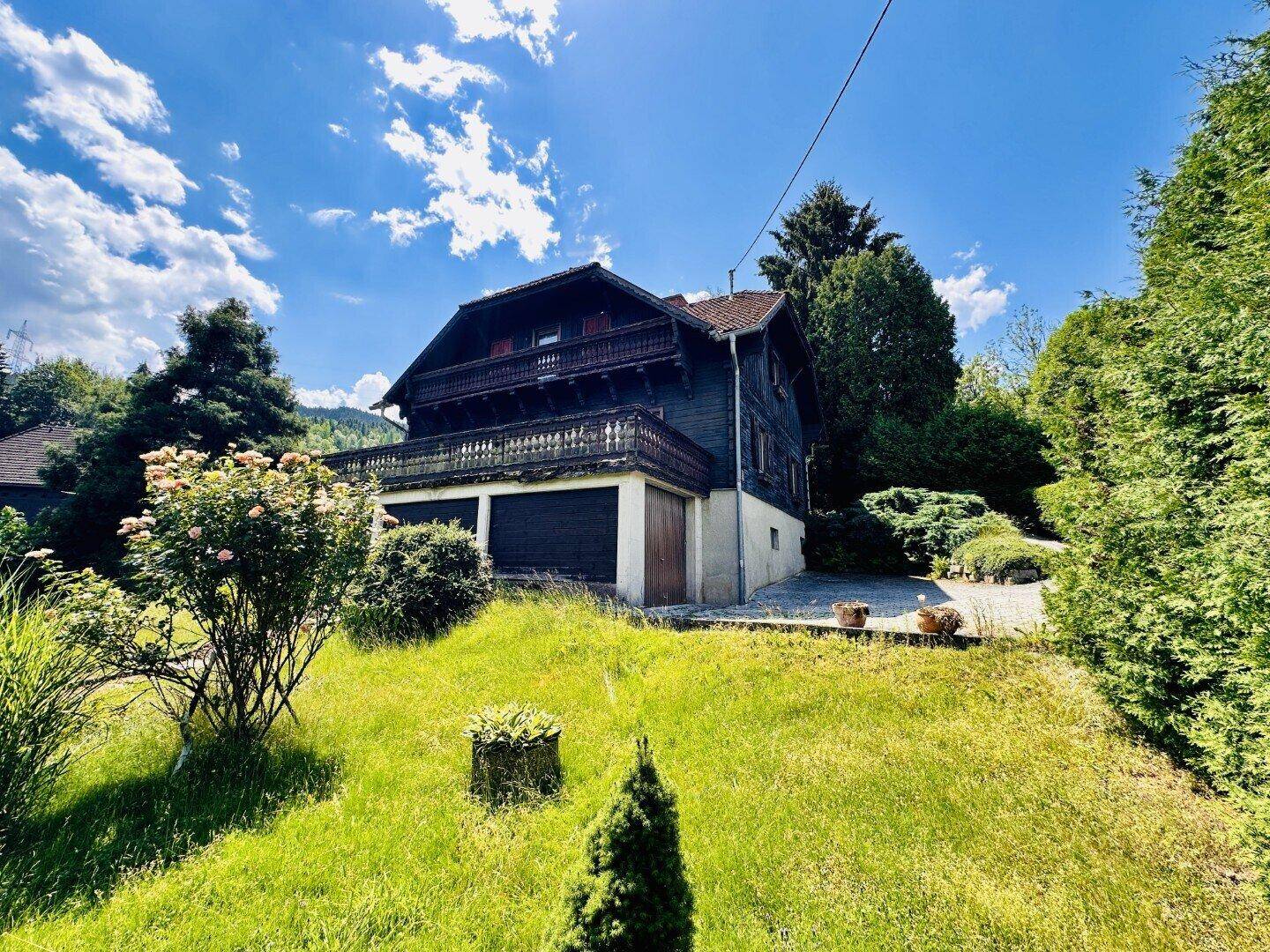 Traditionelles Holzhaus mit Balkon und Garage, umgeben von einem grünen Garten unter blauem Himmel.