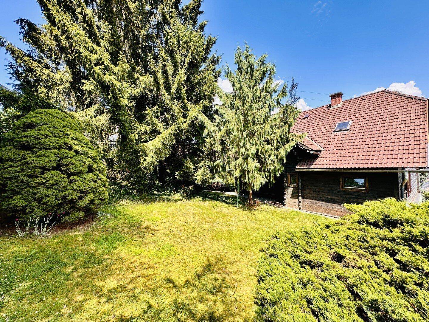 Seitlicher Blick auf ein Haus mit rotem Ziegeldach, umgeben von hohen Bäumen und einer grünen Rasenfläche.