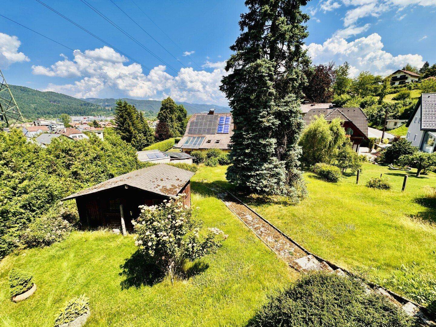 Weitläufiger Garten mit Gartenhaus, hohen Bäumen und Blick auf die umliegende Landschaft und Häuser mit Solaranlagen.