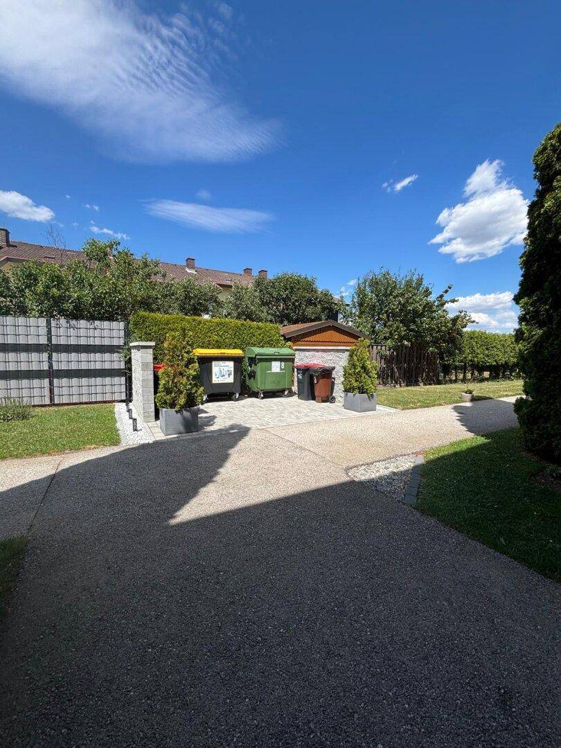 Außenbereich mit Mülltonnen, einem Gehweg und einer Hecke im Hintergrund.