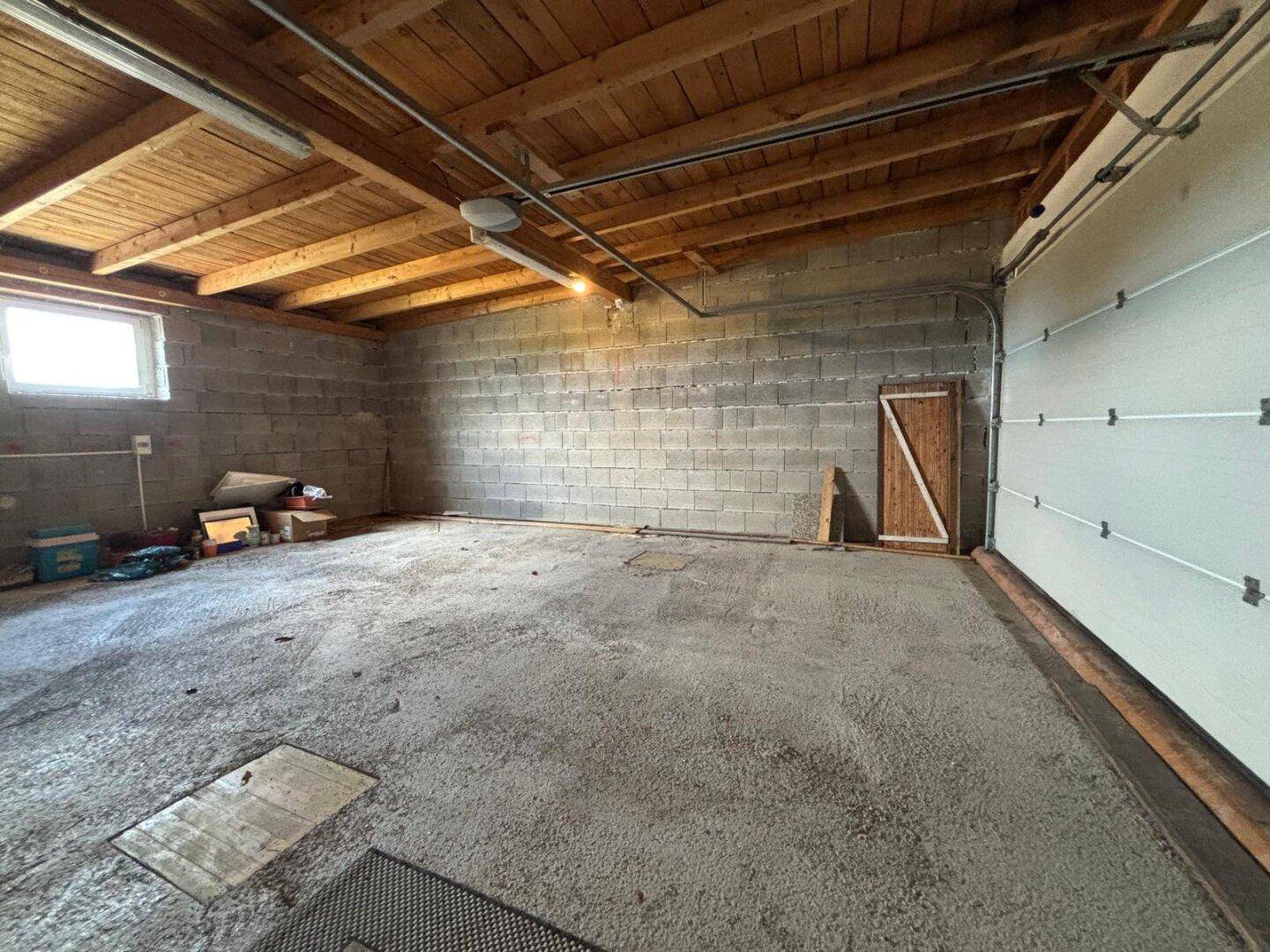 Große Garage mit Betonboden, Holzbalkendecke und Fenster, bietet viel Stauraum.