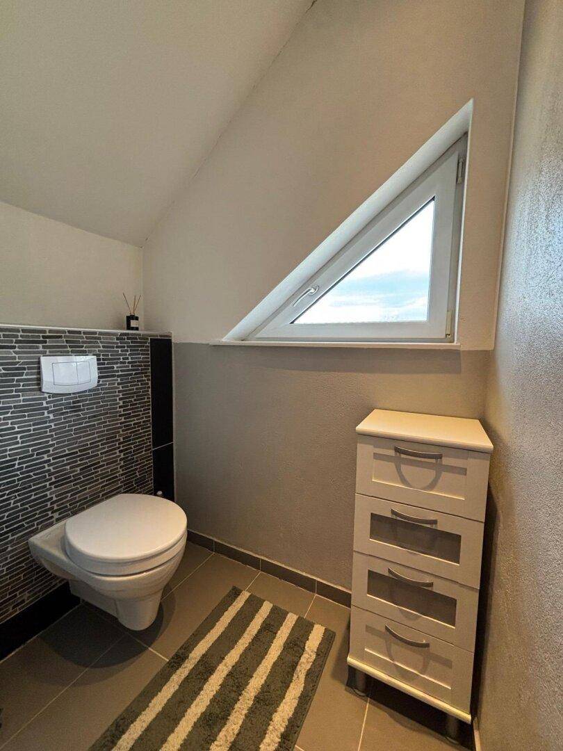 Helles Gäste-WC mit Wand-WC, dreieckigem Fenster und Fliesenboden.