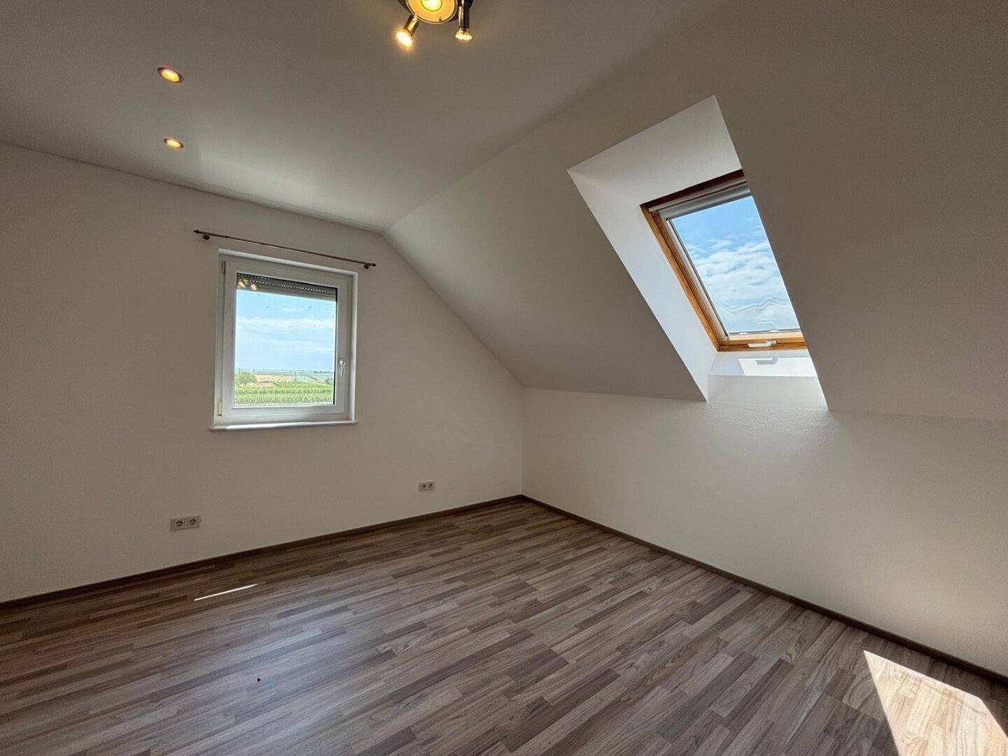 Leeres Zimmer im Dachgeschoss mit Dachfenster, normalem Fenster und Holzboden.