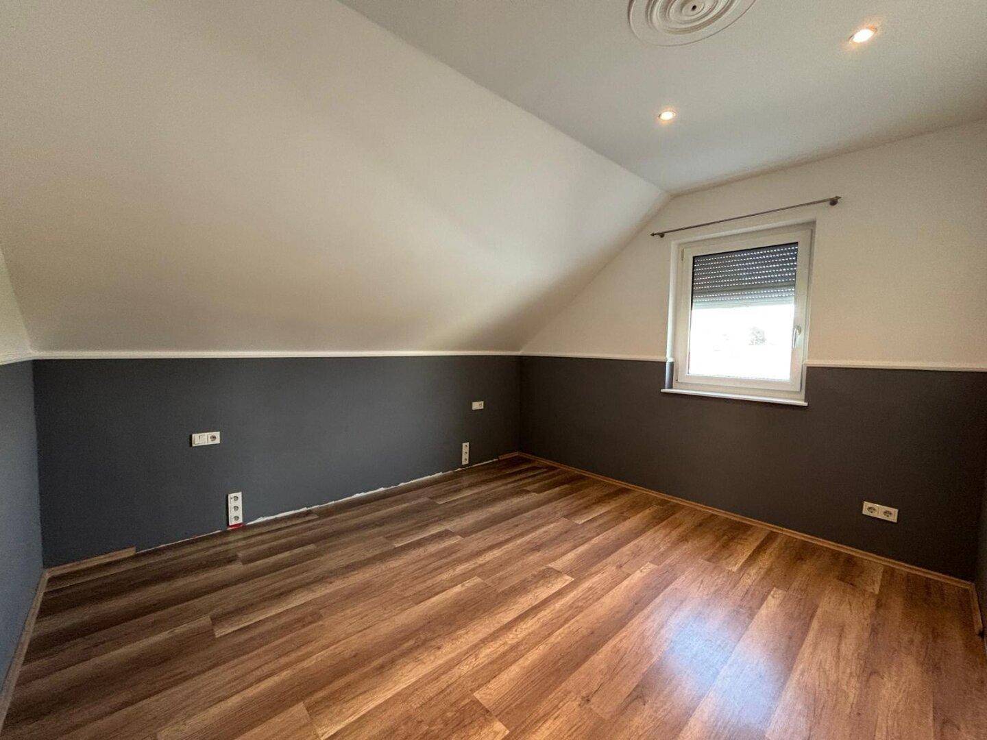 Leeres Schlafzimmer im Dachgeschoss mit Holzboden, Dachschräge und Fenster mit Rollladen.