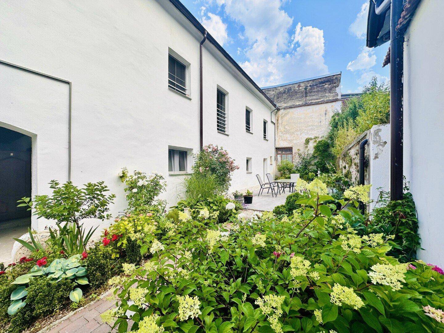 Gepflegter Gartenbereich hinter dem weißen Gebäude mit Sitzgelegenheiten und üppiger Bepflanzung.