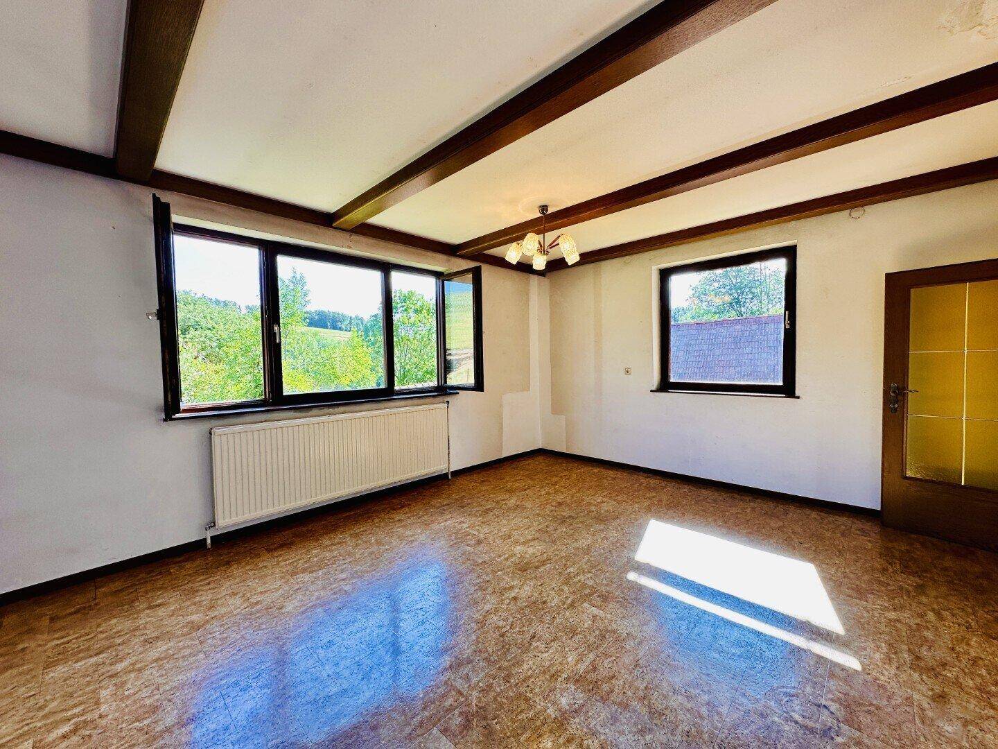 Geräumiges Wohnzimmer mit Holzbalkendecke, großen Fenstern und Blick ins Grüne.