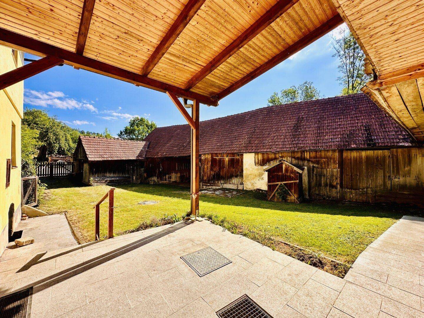 Überdachte Terrasse mit Holzdach und Blick auf den Rasen und die Nebengebäude.