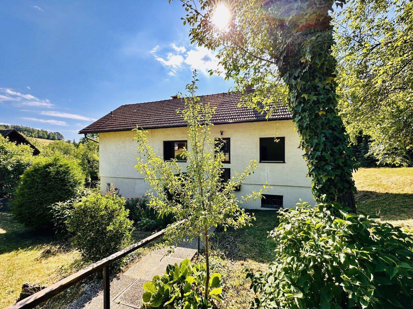 Hausfassade mit weißem Putz und Ziegeldach, umgeben von Bäumen und Sträuchern an einem sonnigen Tag.