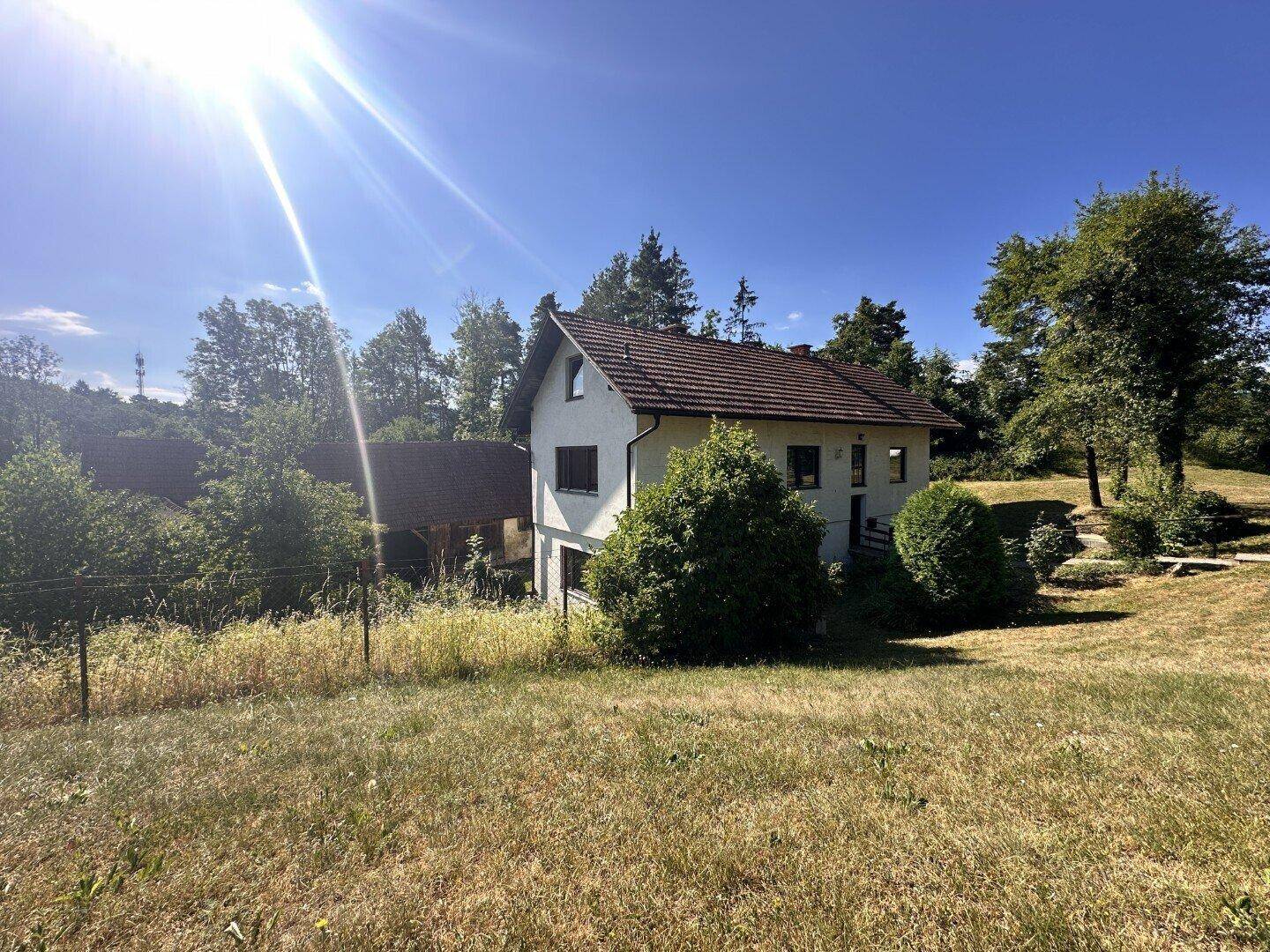 Haus in ländlicher Umgebung mit weißer Fassade und Ziegeldach, umgeben von Wiesen und Bäumen.