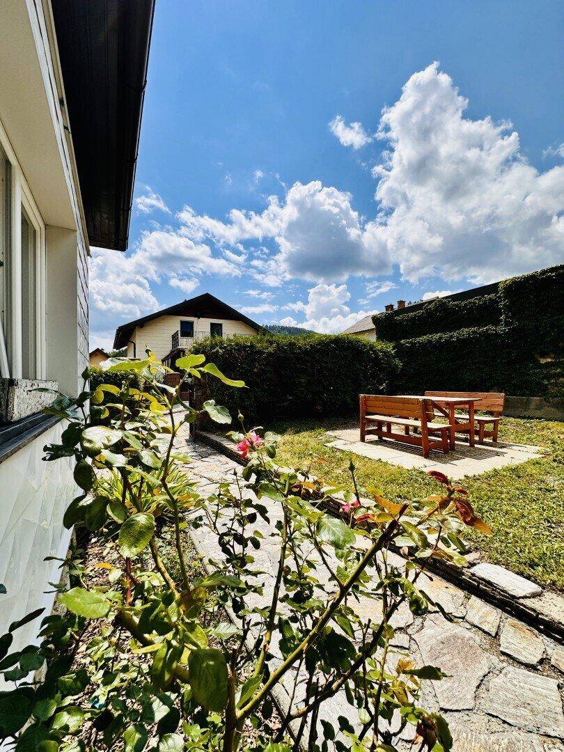 Sonniger Gartenbereich mit Rasen, Holztisch und Bänken, umgeben von Hecken.
