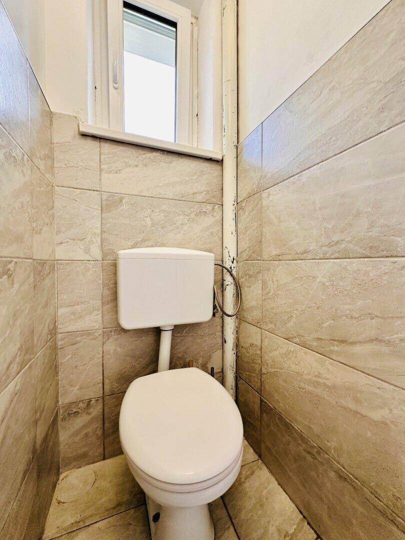 Separate Toilette mit beigefarbenen Fliesen, Fenster und einfacher Ausstattung.