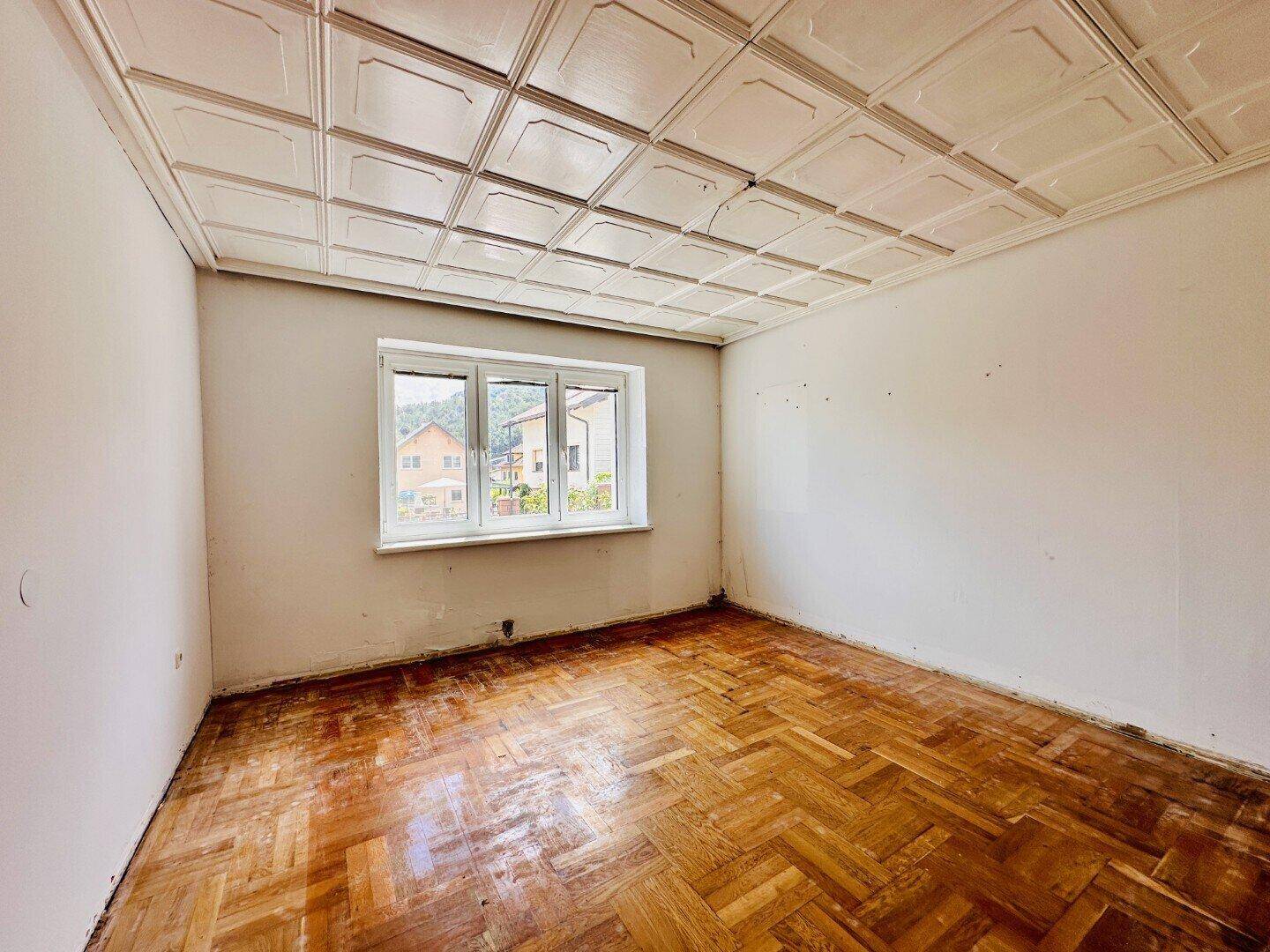 Leerer Raum mit Fischgrätparkett, weißer Decke und Fenster mit Blick auf die Umgebung.