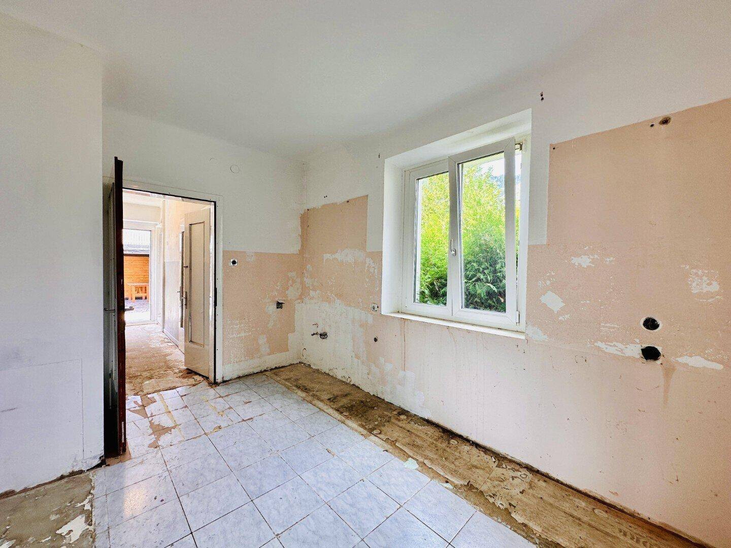 Leere Küche mit Fliesenboden, Renovierungsbedarf und Fenster mit Blick in den Garten.