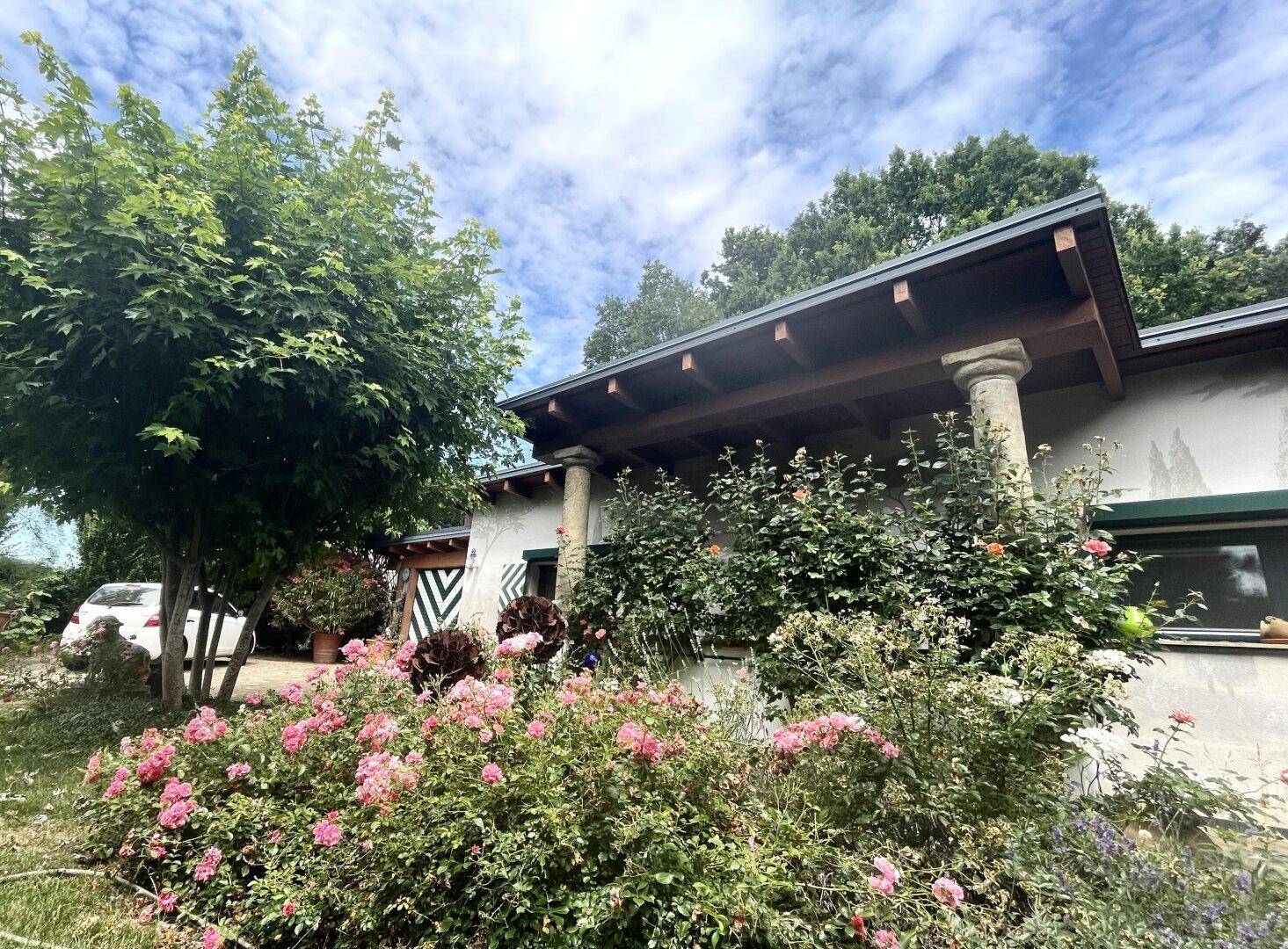 Hausfassade mit rustikalen Säulen, umgeben von blühenden Pflanzen und einem gepflegten Gartenbereich.