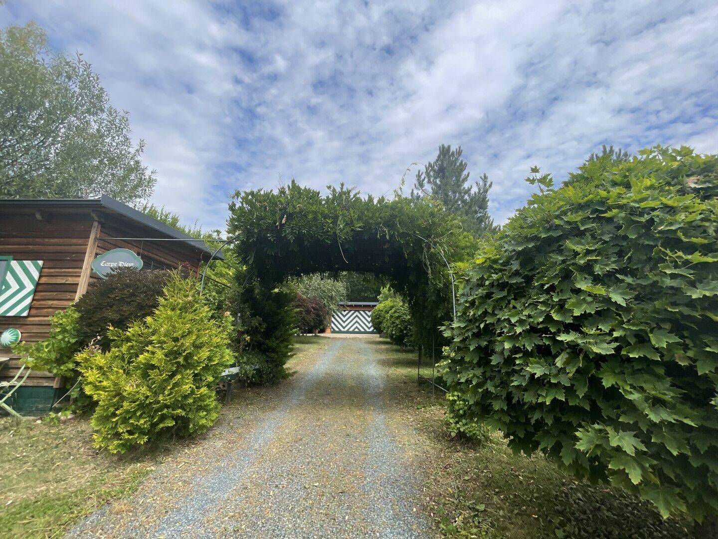 Zufahrt zum Anwesen, gesäumt von üppigem Grün und einem begrünten Torbogen unter blauem Himmel.