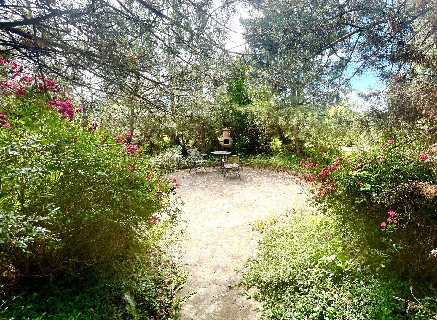 Gemütlicher Gartenbereich mit Steinterrasse, Sitzgelegenheiten und einem Grillkamin umgeben von Bäumen.