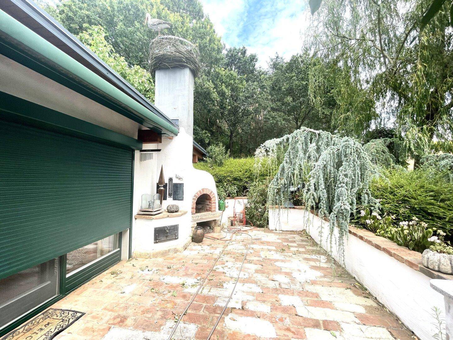 Terrasse mit Ziegelboden, Außenkamin und grünen Rollläden, umgeben von Bäumen.