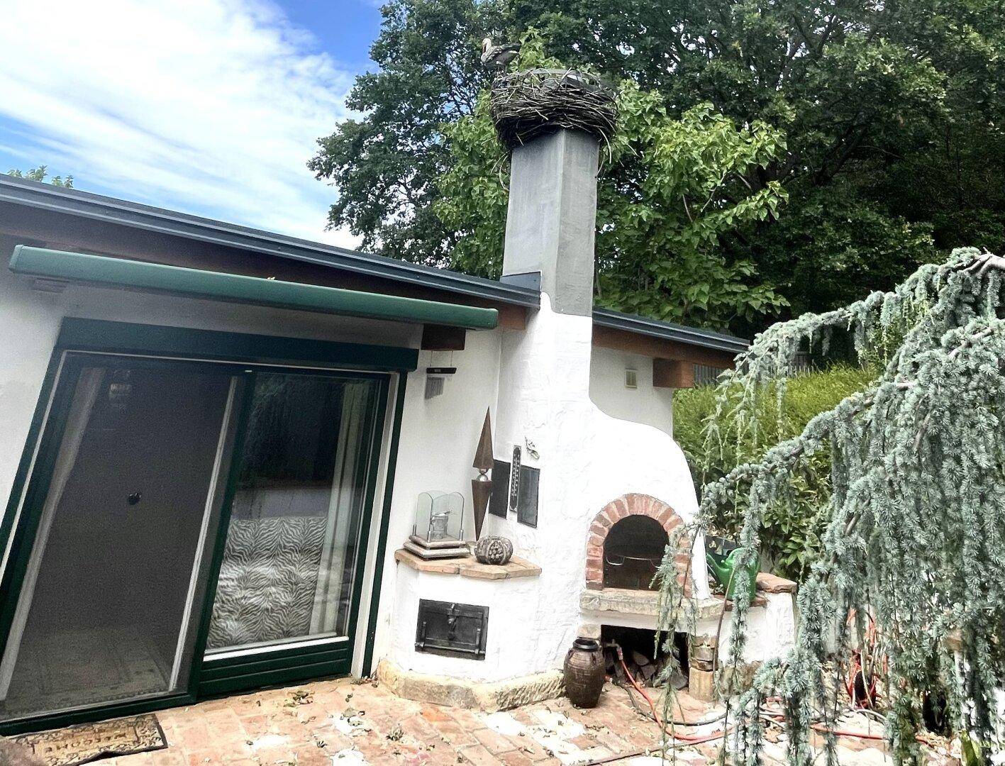 Hausfassade mit Außenkamin, grünen Rollläden und einer Terrasse, umgeben von üppiger Vegetation.