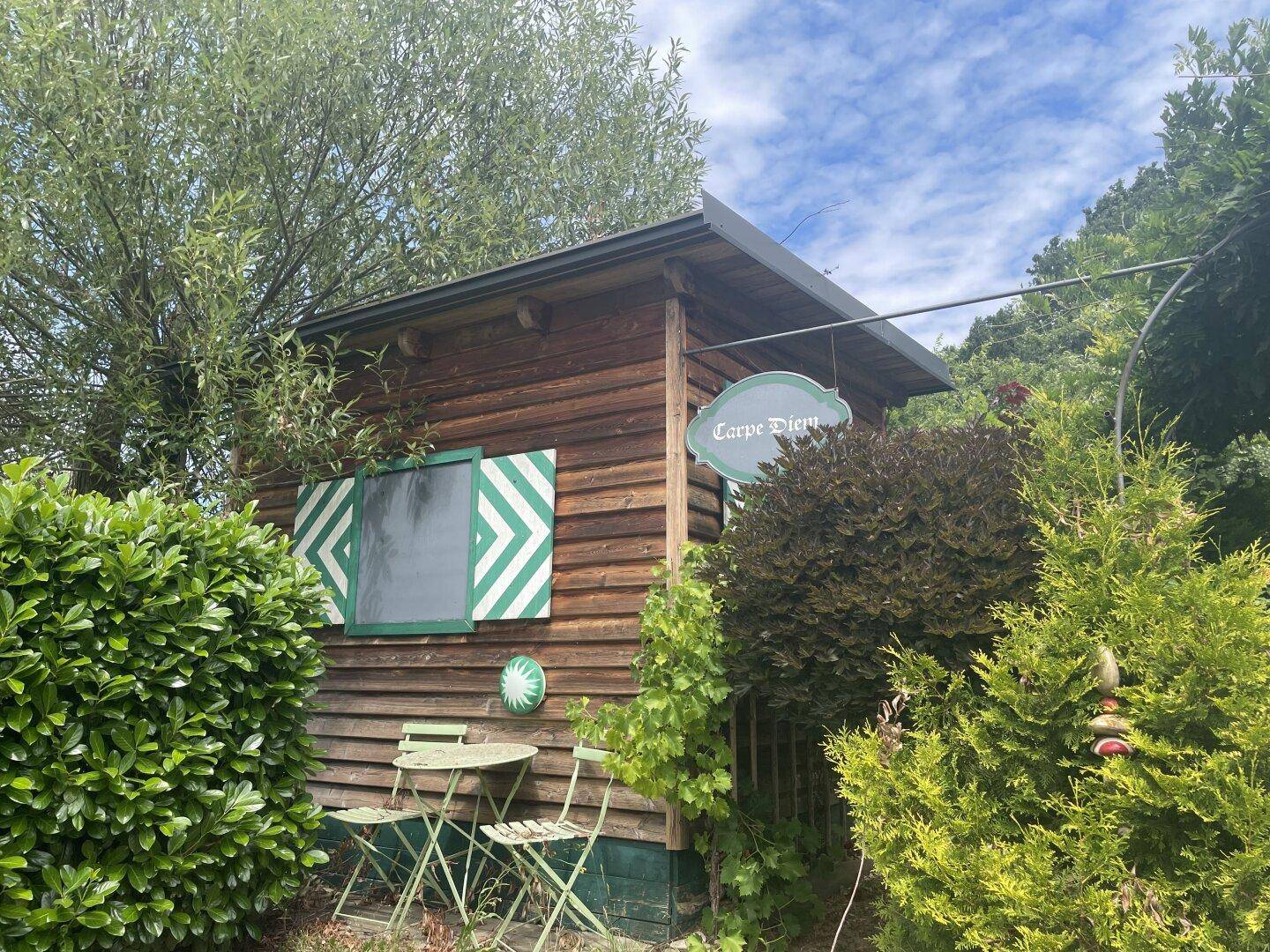 Kleines Gartenhaus aus Holz mit Sitzgelegenheiten und grünen Fensterläden, umgeben von Pflanzen.