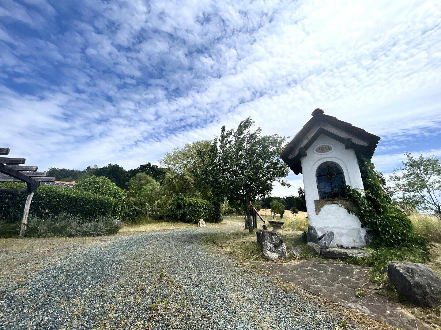Ländlicher Weg mit Schotter und einer kleinen Kapelle unter blauem Himmel.