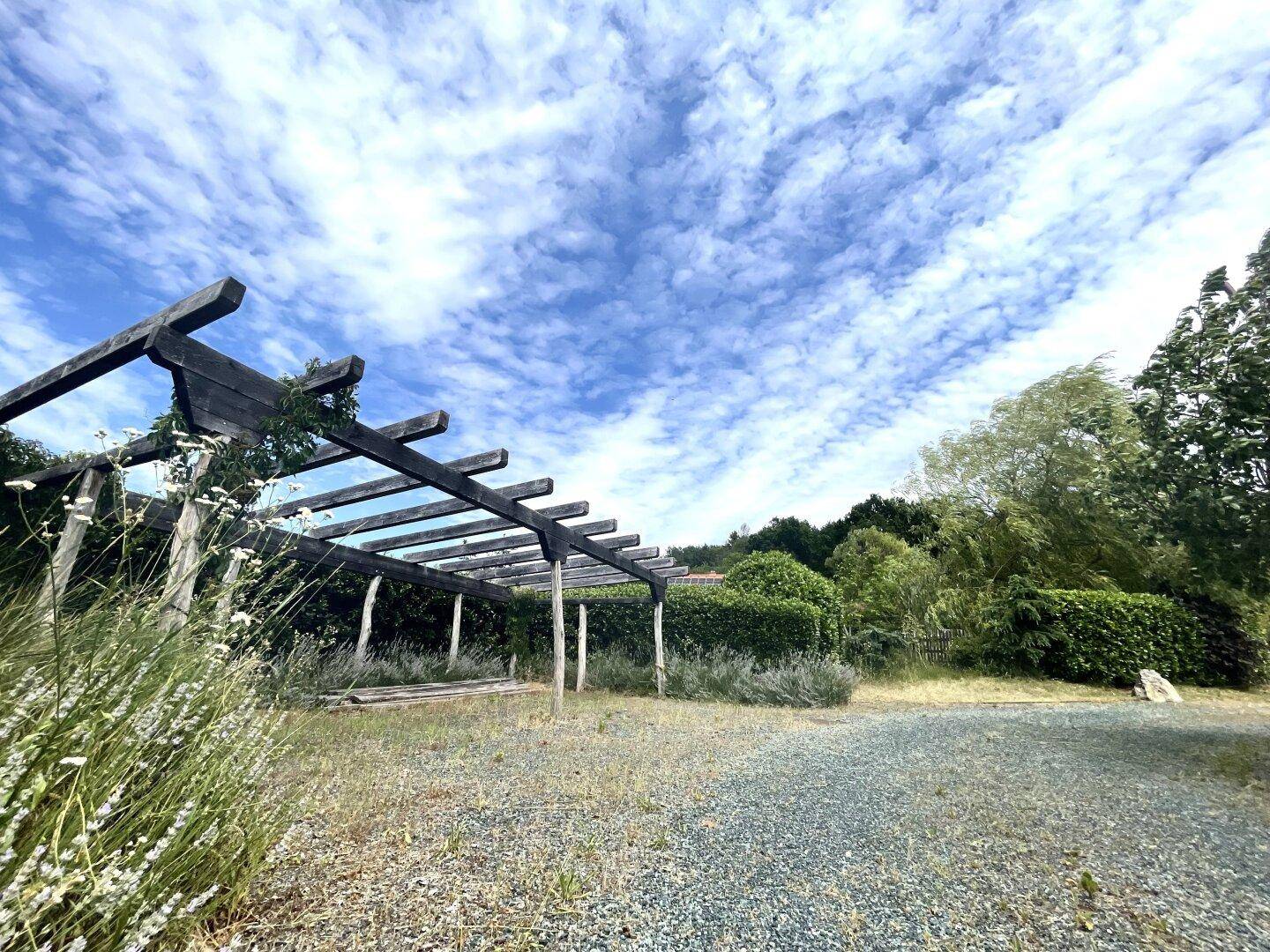Hölzerne Pergola in einem Kiesbereich, umgeben von Pflanzen unter weitem Himmel.