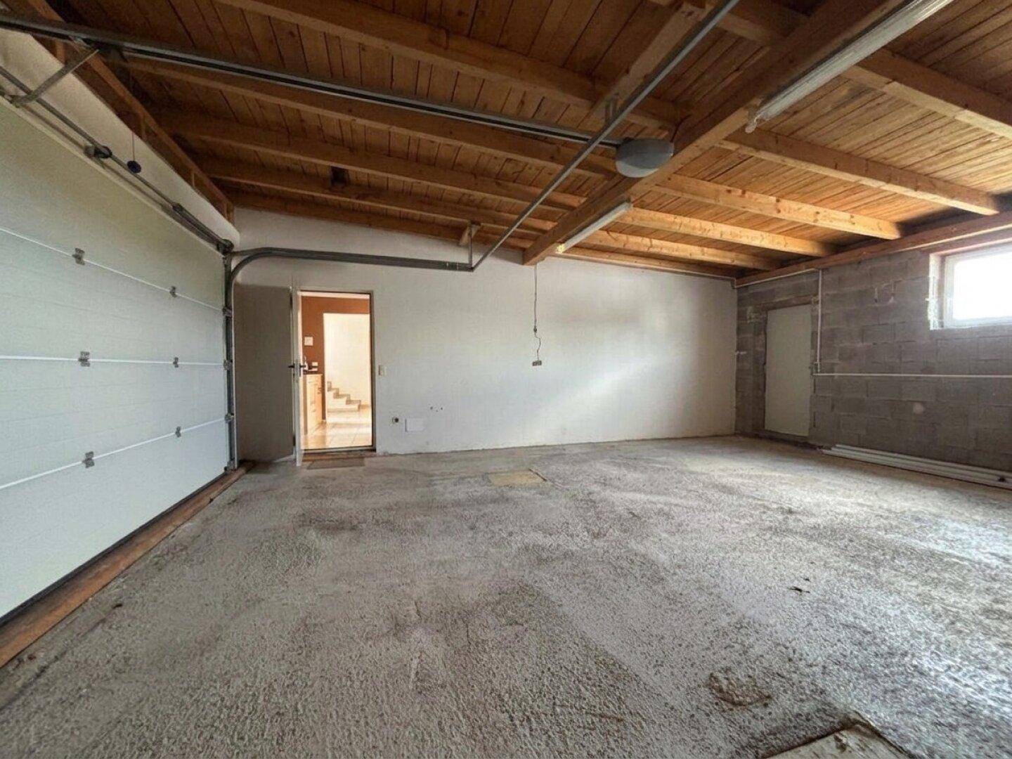 Geräumige Garage mit Betonboden und Holzbalkendecke, mit Zugang zu weiteren Räumen.