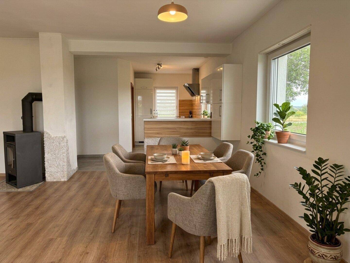 Essbereich mit Holztisch, Kaminofen und offenem Übergang zur Küche, mit Blick ins Grüne.