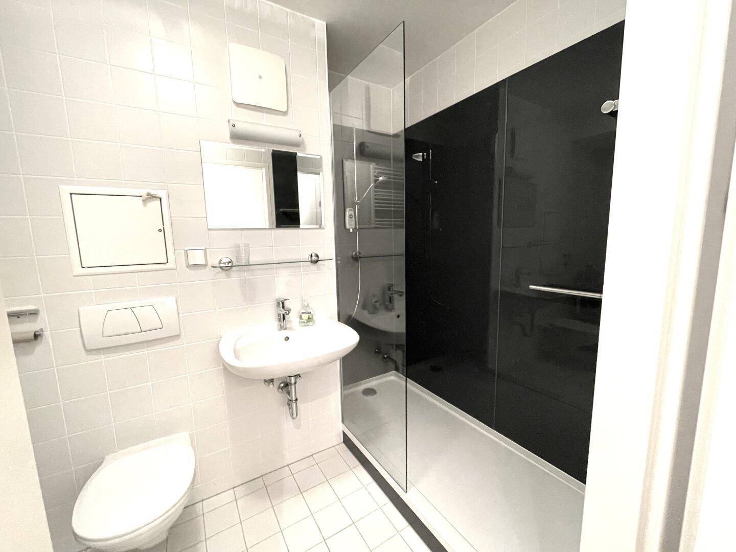 Helles Badezimmer mit weißen Fliesen, WC, Waschbecken und Duschbereich.