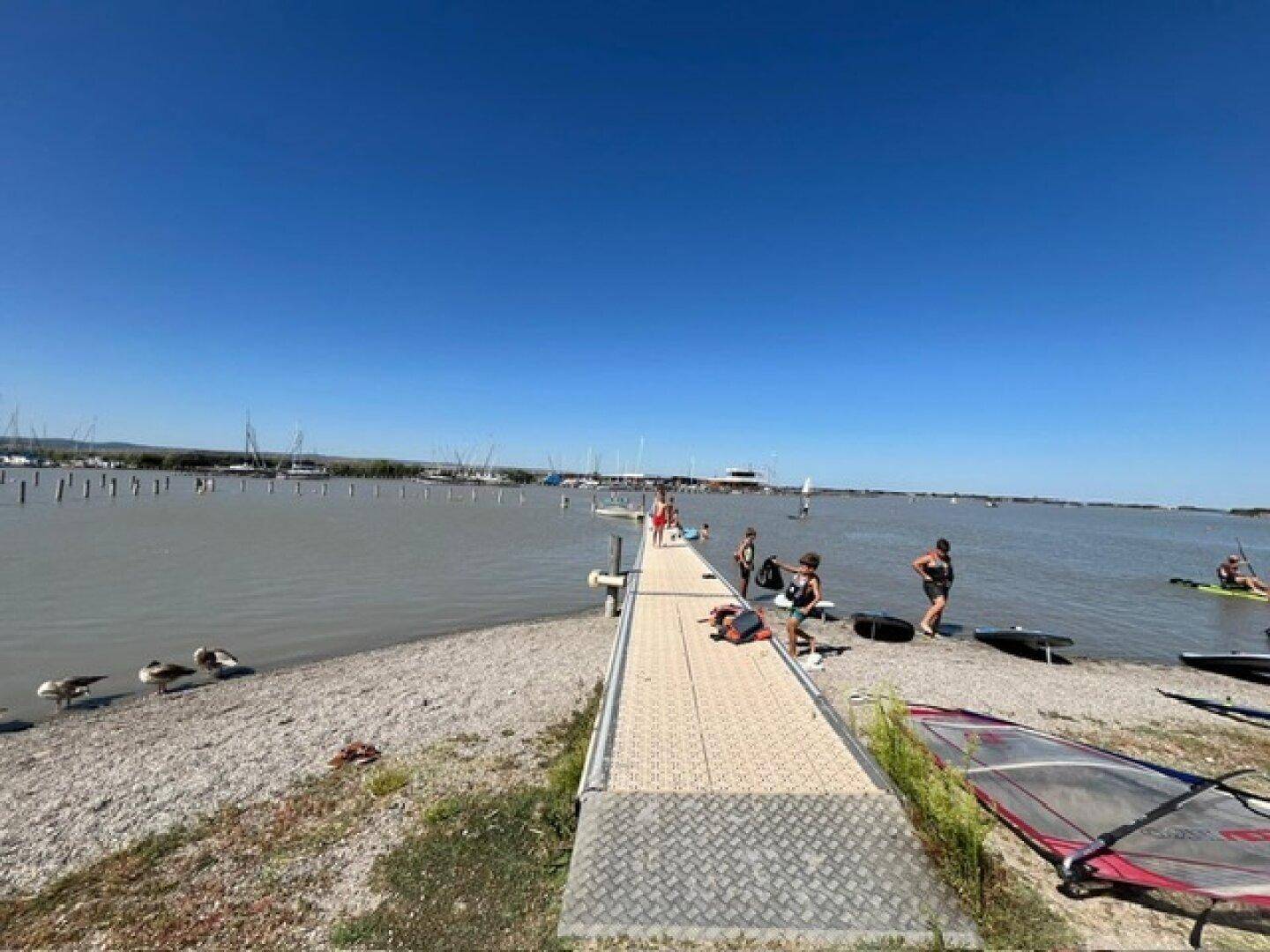 Strandbereich mit einem Steg, Menschen am Wasser und Segelbooten im Hintergrund unter blauem Himmel.