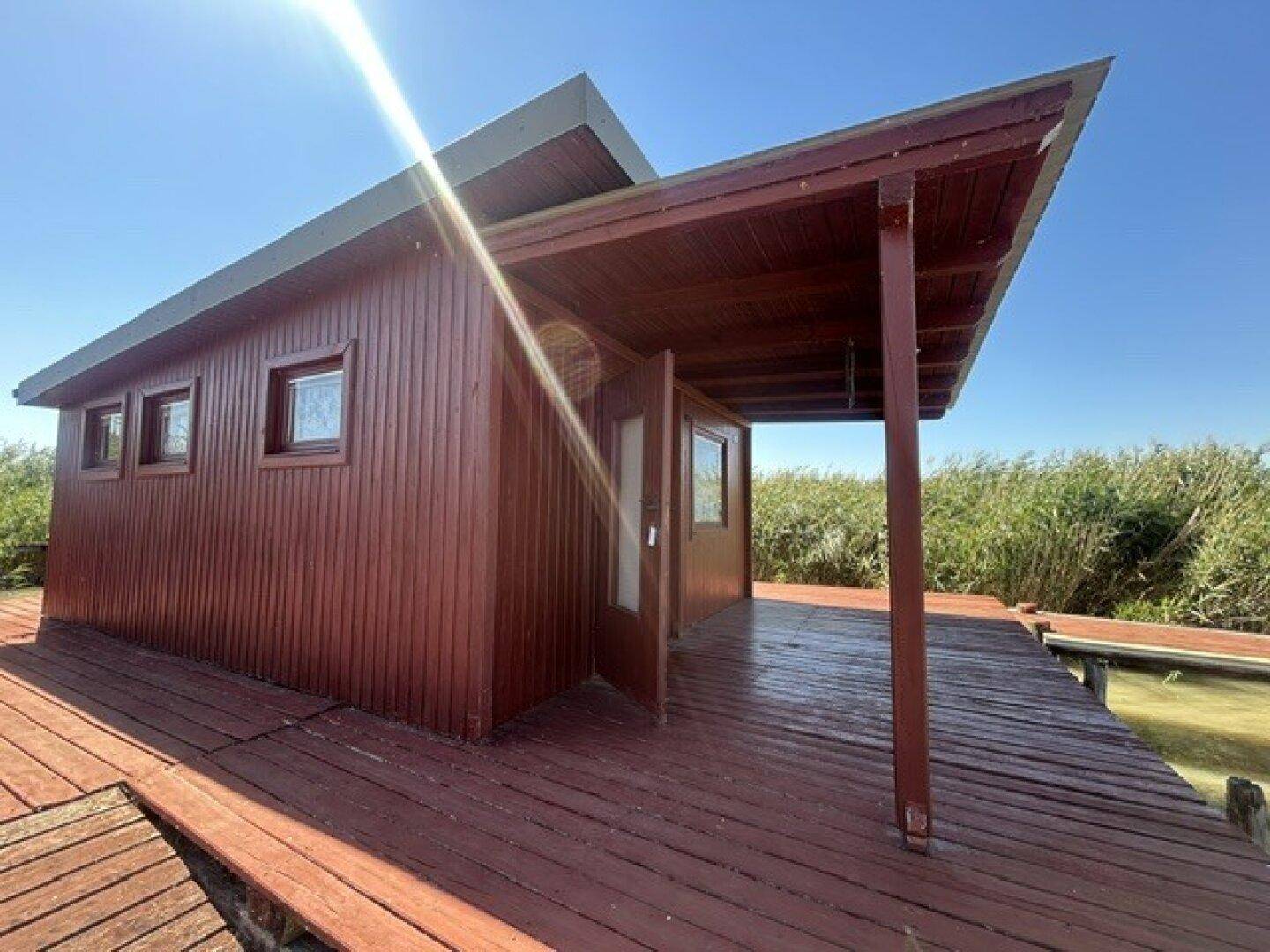 Seitliche Ansicht der Holzhütte mit breiter Holzterrasse und Blick auf das Wasser unter blauem Himmel.