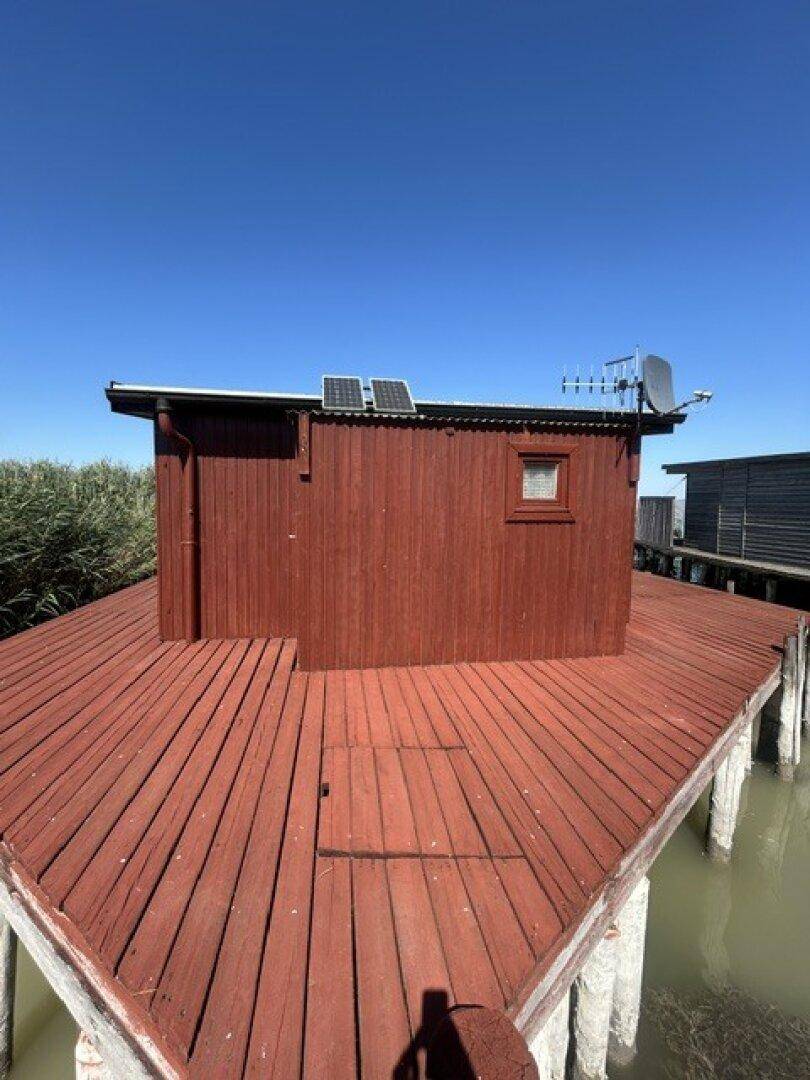 Rückseite der Holzhütte mit Solarpaneelen auf dem Dach und einer weitläufigen Holzplattform über dem Wasser.