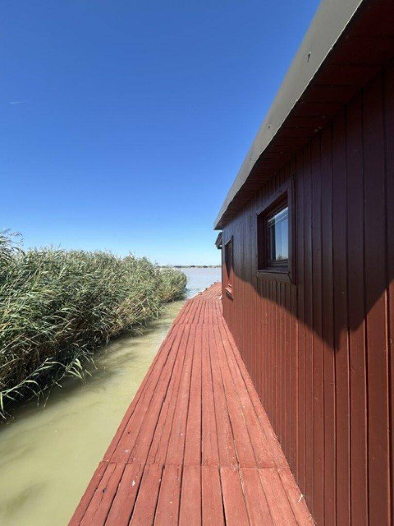 Langer Holzsteg entlang der Holzhütte mit Schilf am Ufer und weitem Blick über das Wasser.