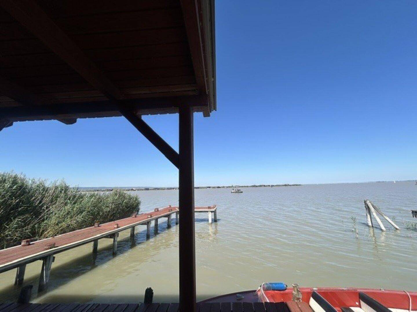 Blick von der überdachten Holzterrasse auf den See mit einem langen Holzsteg und klarem Himmel.