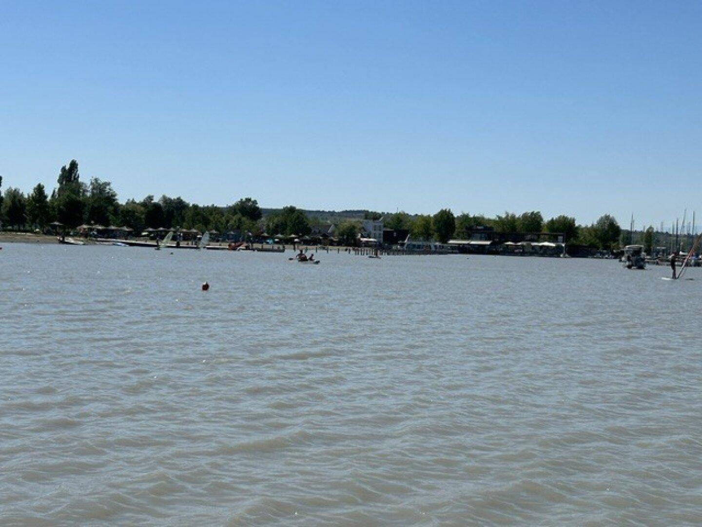 Weite Wasserfläche eines Sees mit zahlreichen Booten und der grünen Uferlinie in der Ferne.
