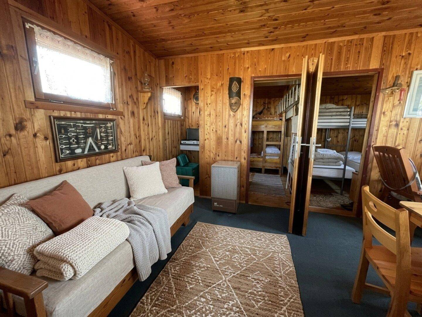 Gemütliches Wohnzimmer mit holzvertäfelten Wänden, Sofa und Blick in ein Schlafzimmer mit Etagenbetten.