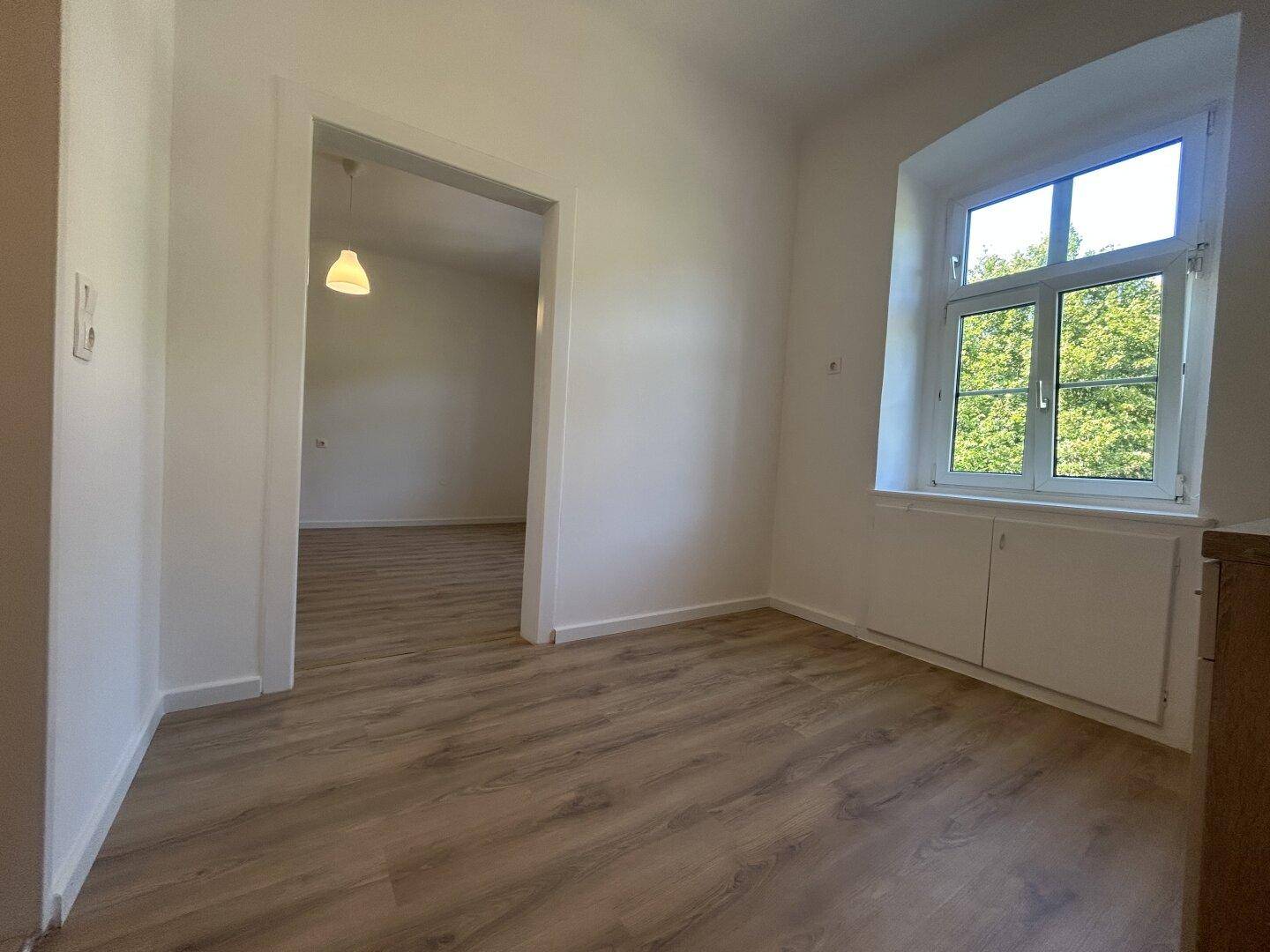 Heller, leerer Raum mit Laminatboden und einem Fenster, das viel Tageslicht hereinlässt.