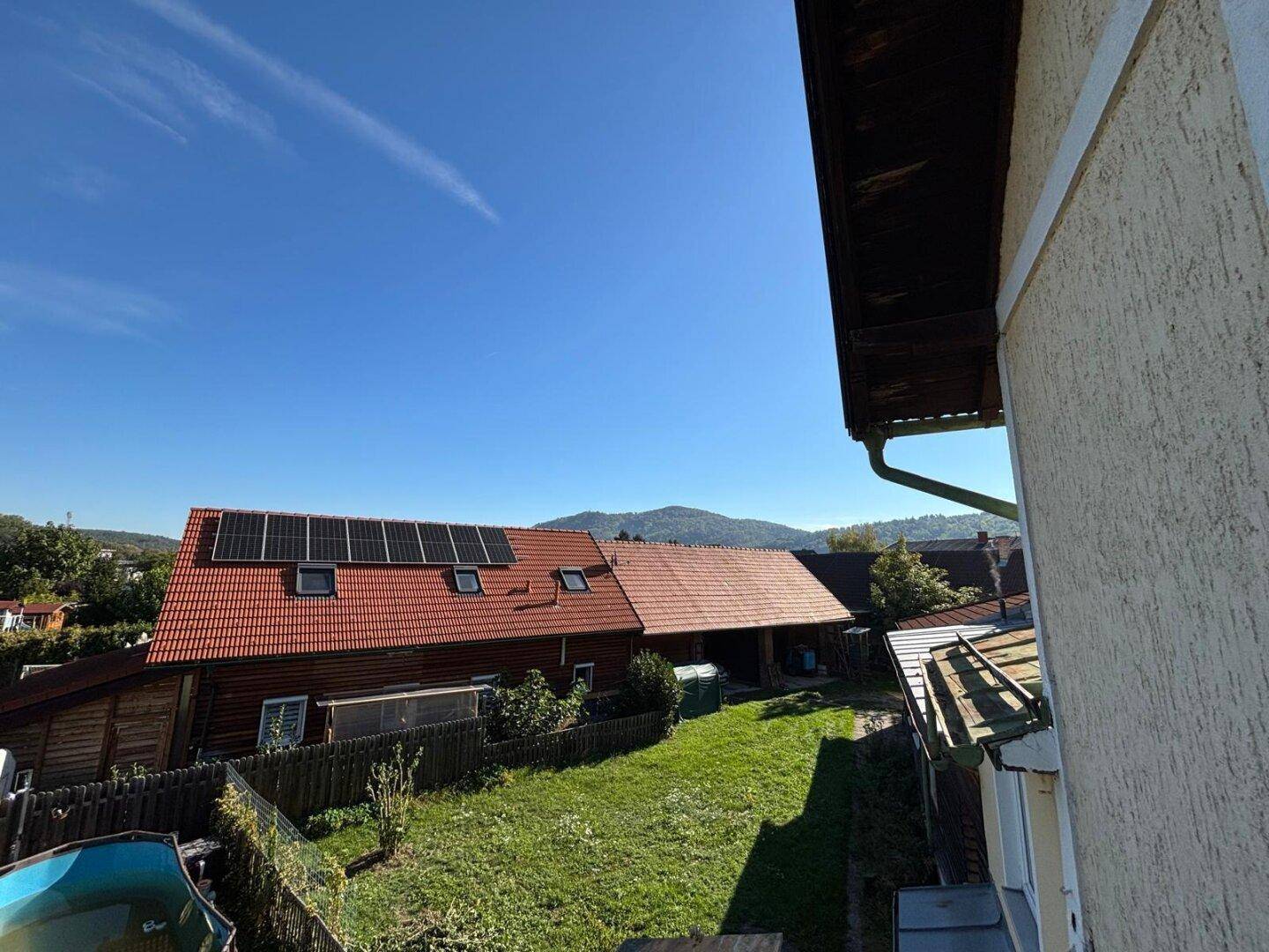 Gartenbereich mit Rasen und Bäumen, umgeben von Häusern mit roten Dächern und Bergblick.