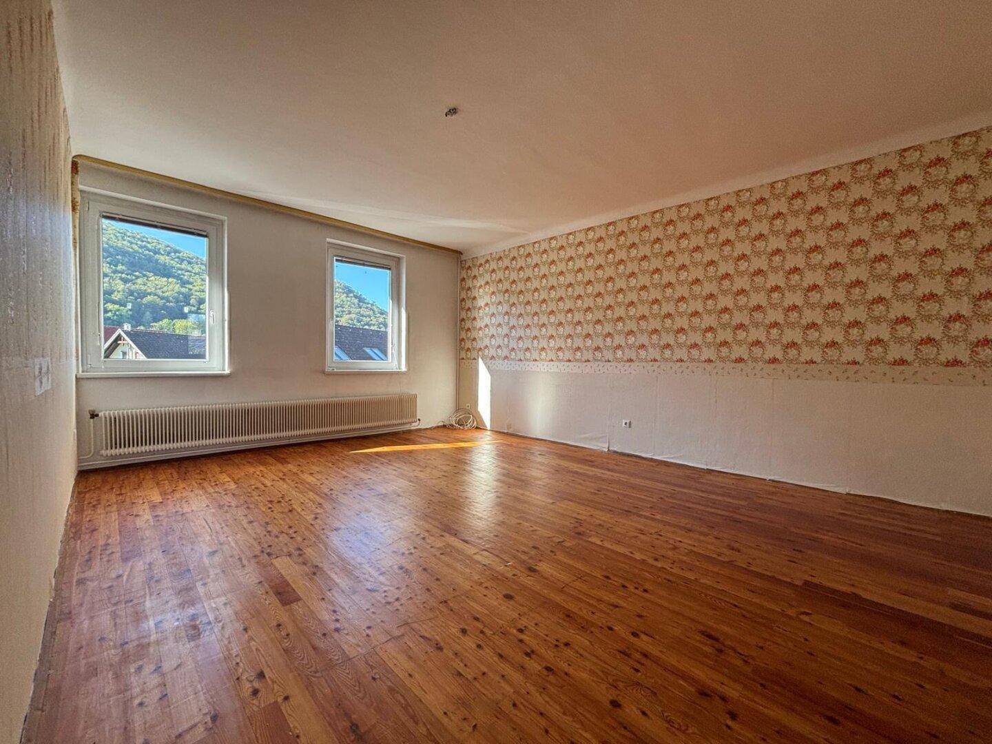 Helles, leeres Zimmer mit Holzboden, gemusterter Tapete und Blick auf grüne Hügel.