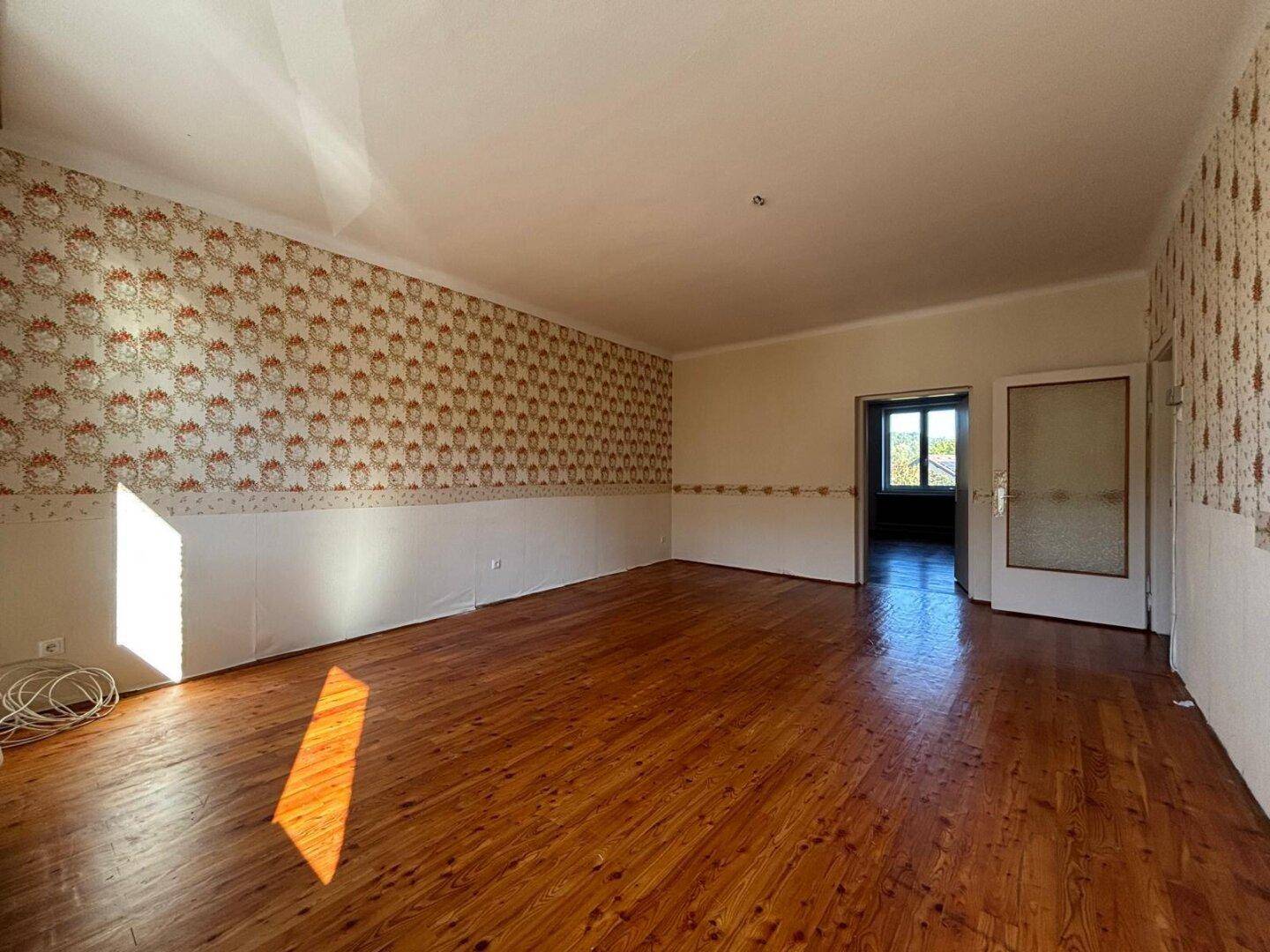Geräumiges Zimmer mit Holzboden und gemusterter Tapete, einladender Durchgang zu einem weiteren Raum.